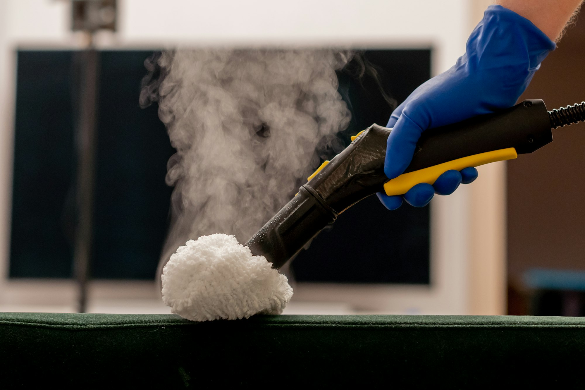apartment cleaner in yellow apron does a steam cleaning of a green sofa with professional equipment